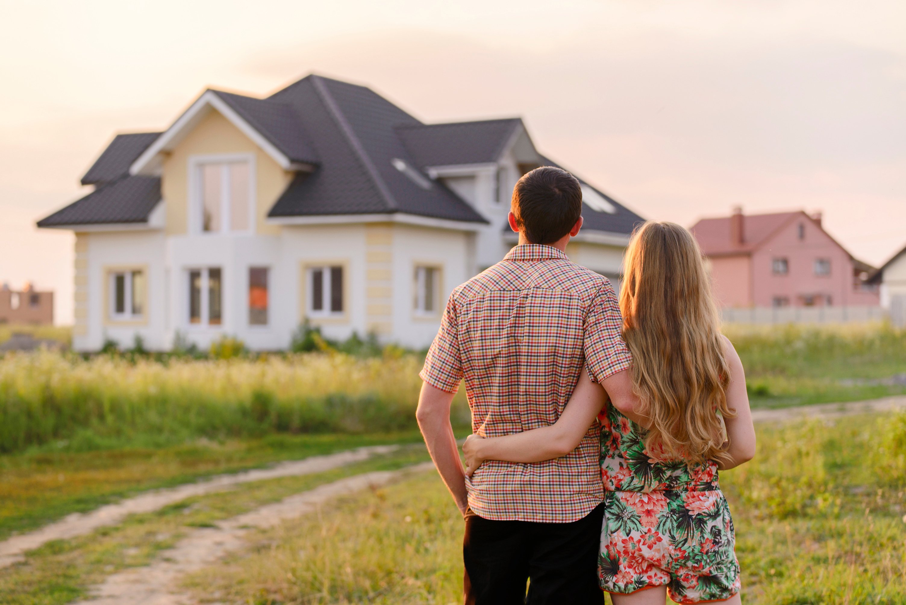 A couple stands arm in arm, looking at a modern two-story house with a dark roof and light-colored exterior, surrounded by grass and a dirt path, during sunset.