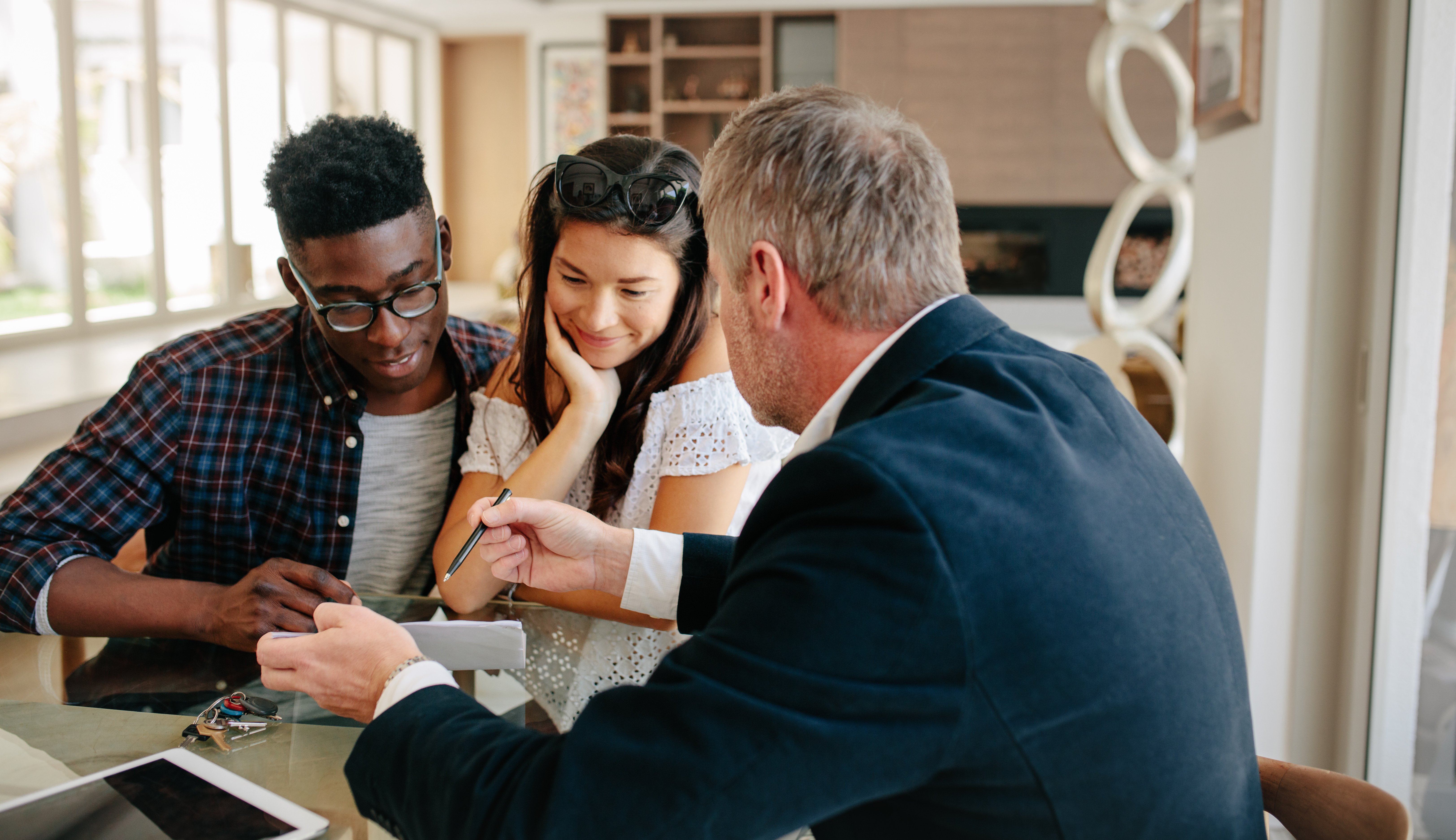 A couple sits at a table with a professional advisor, reviewing documents together. The man in the plaid shirt and glasses leans in while the woman in a white blouse smiles thoughtfully. The advisor, dressed in a suit, gestures with a pen toward the paperwork. A tablet and a set of keys are on the table.