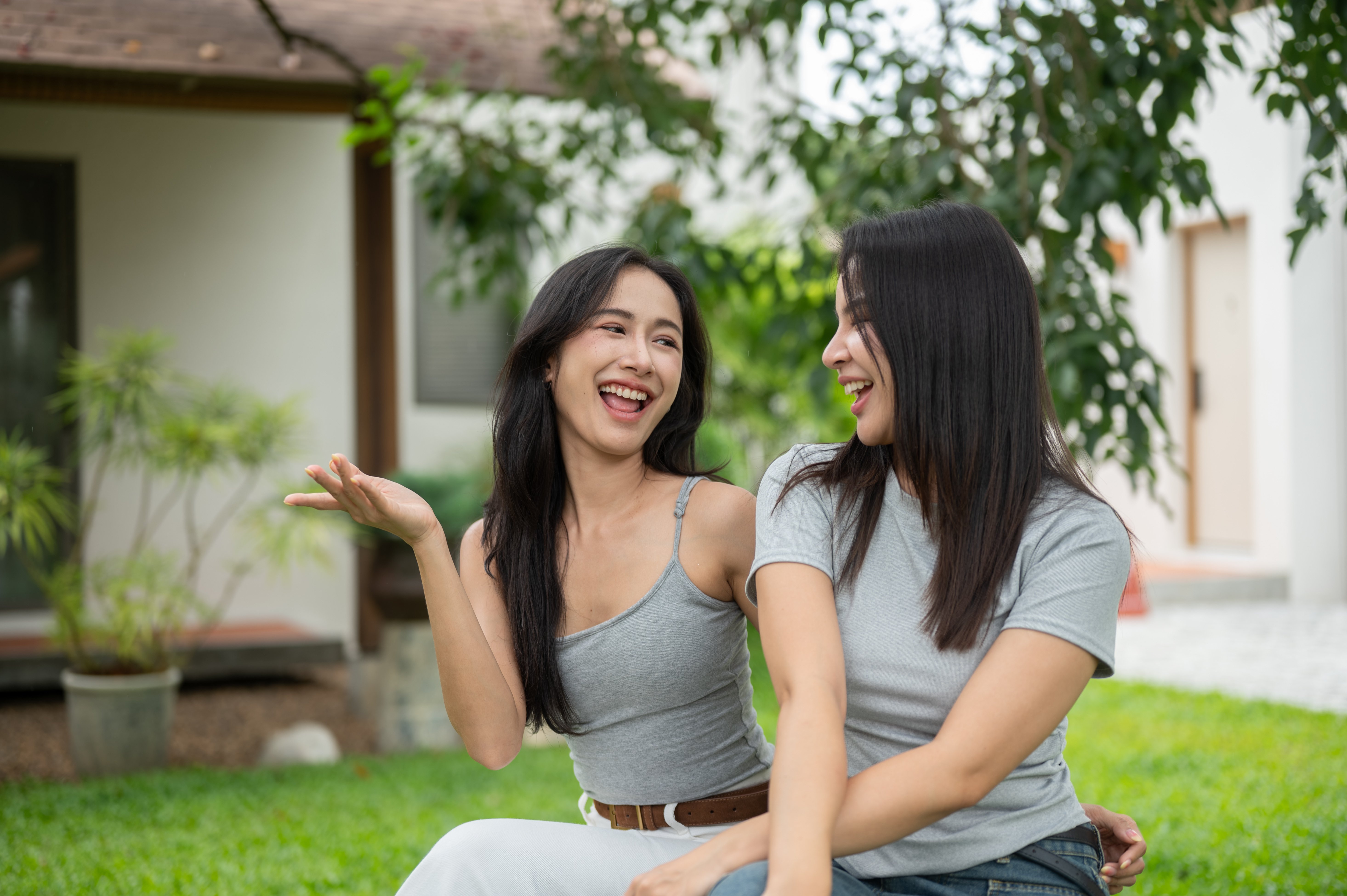 Two female friends chatting and laughing together on the green grass lawn in front of a house.
