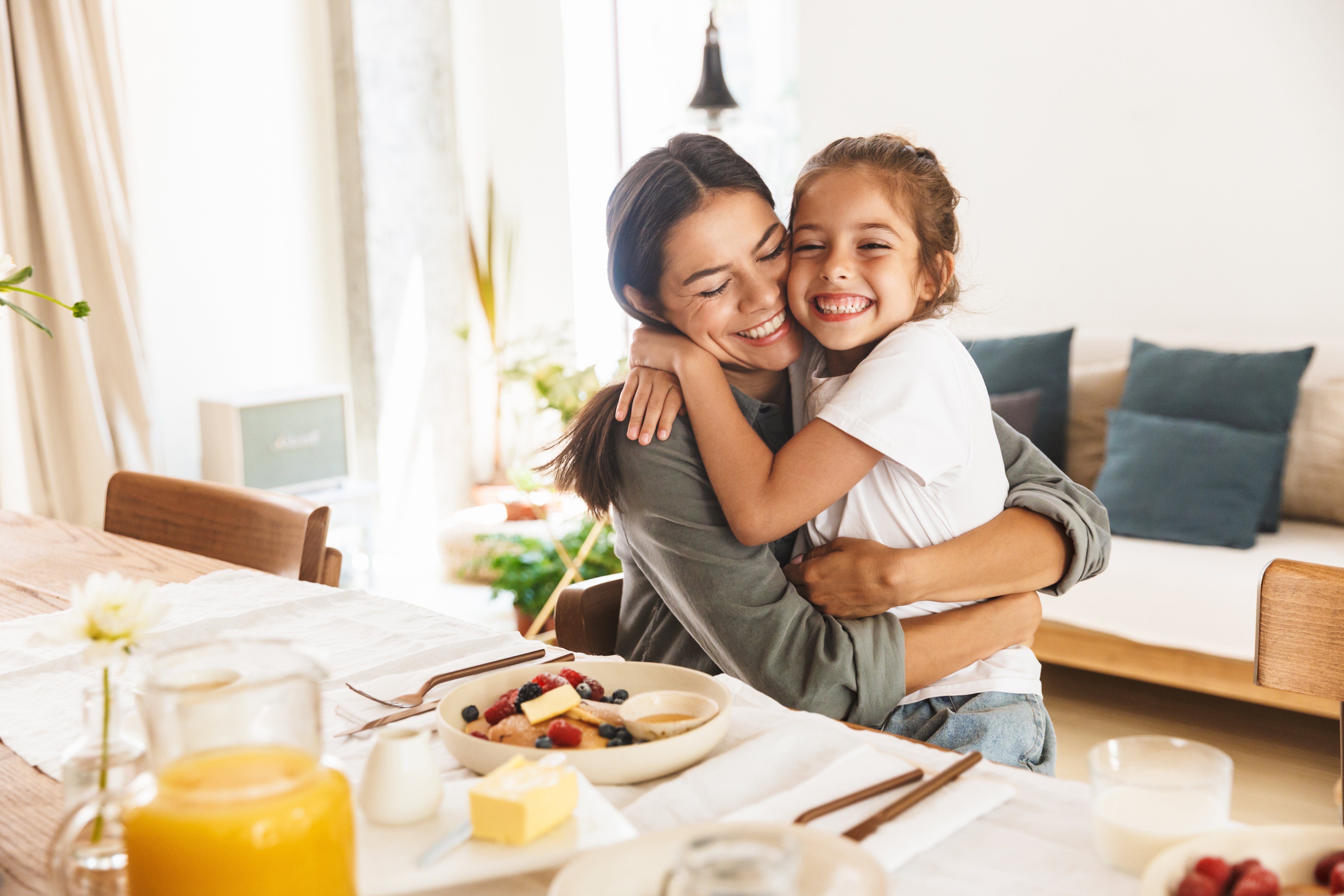Image of happy family mother and little daughter hugging while having breakfast at home in morning.