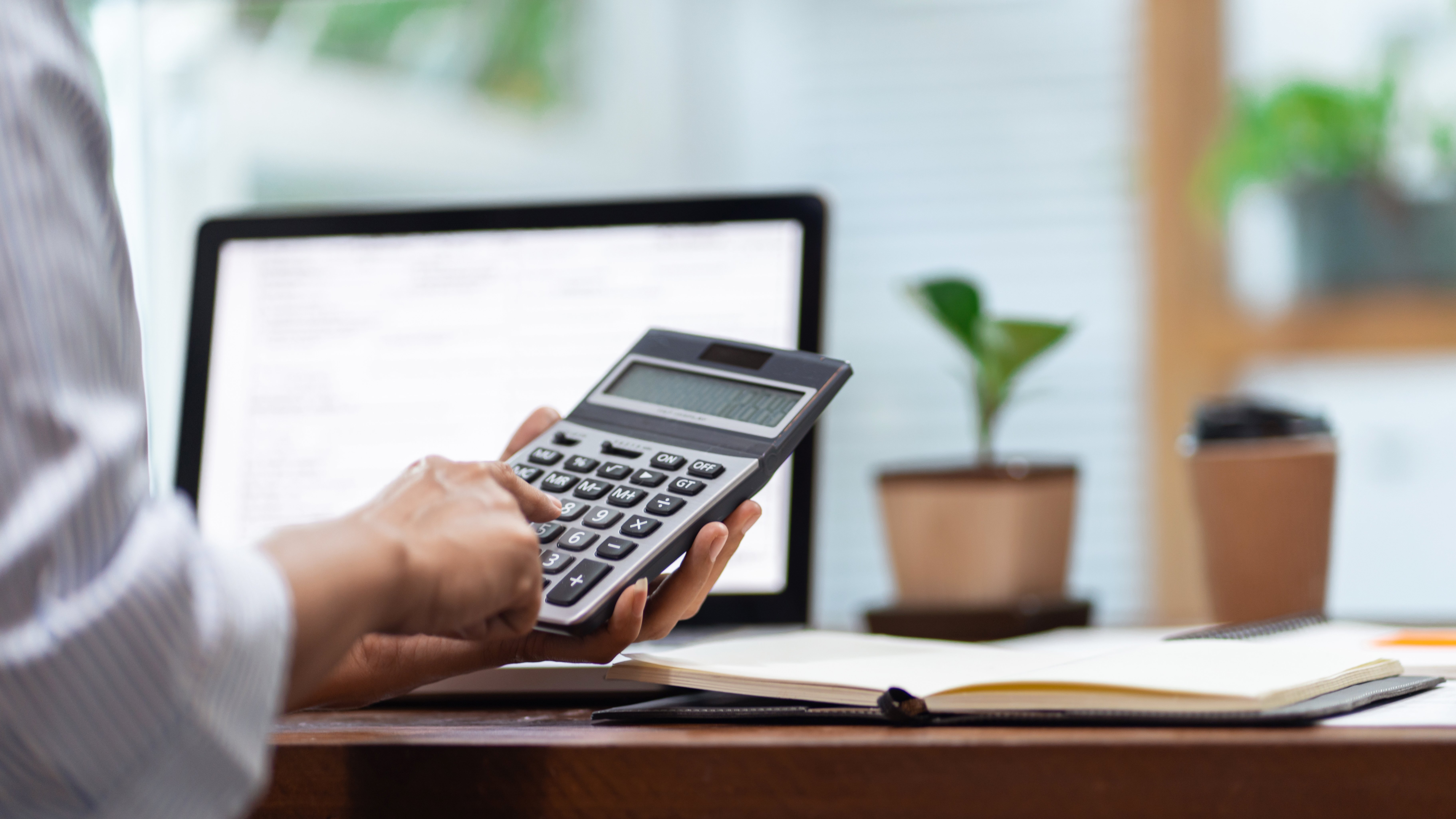 Person using a large calculator at a desk with an open notebook, laptop, coffee cup, and potted plant in the background.