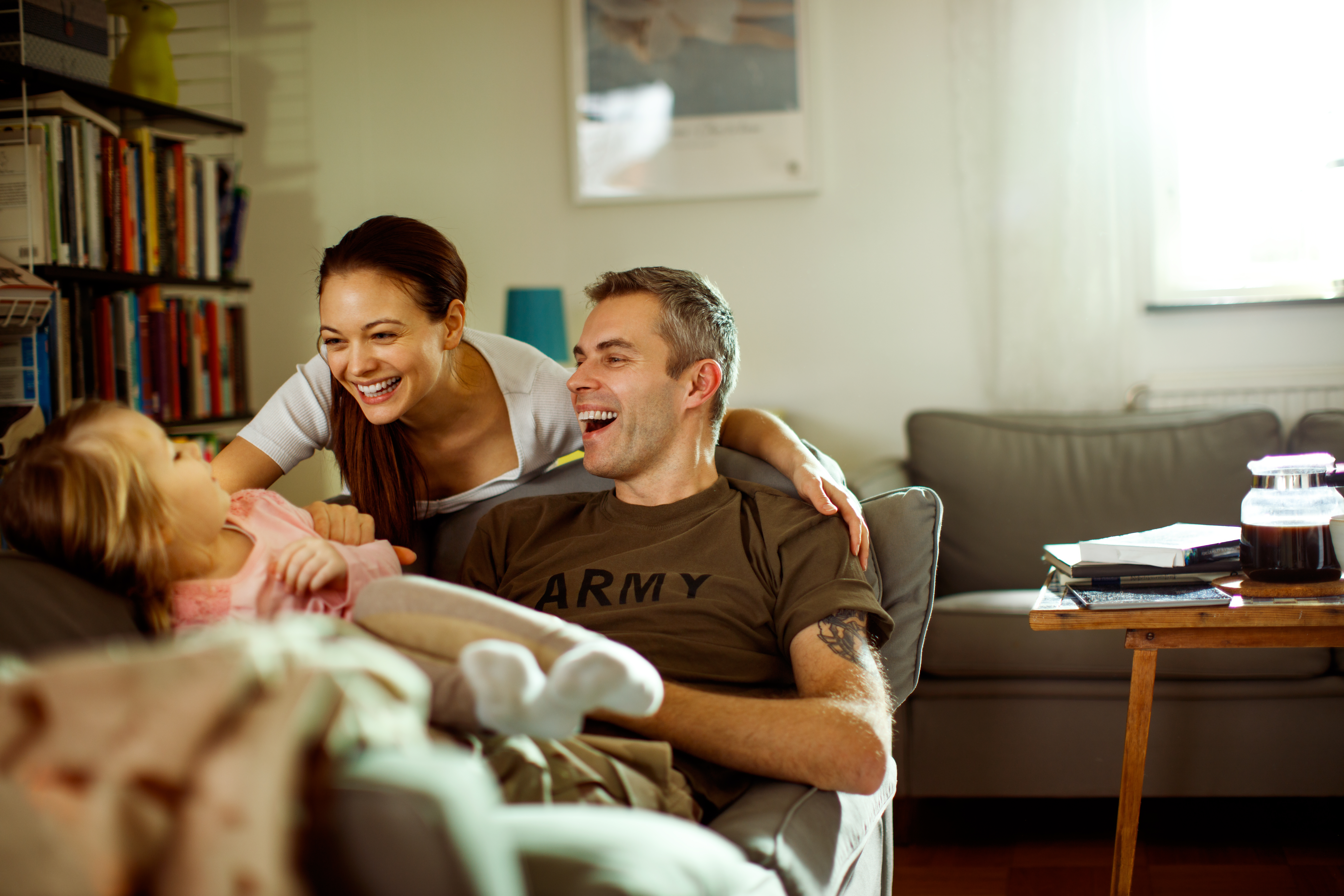 A young couple smile and laugh with their daughter. The man is wearing a brown shirt that says "Army."