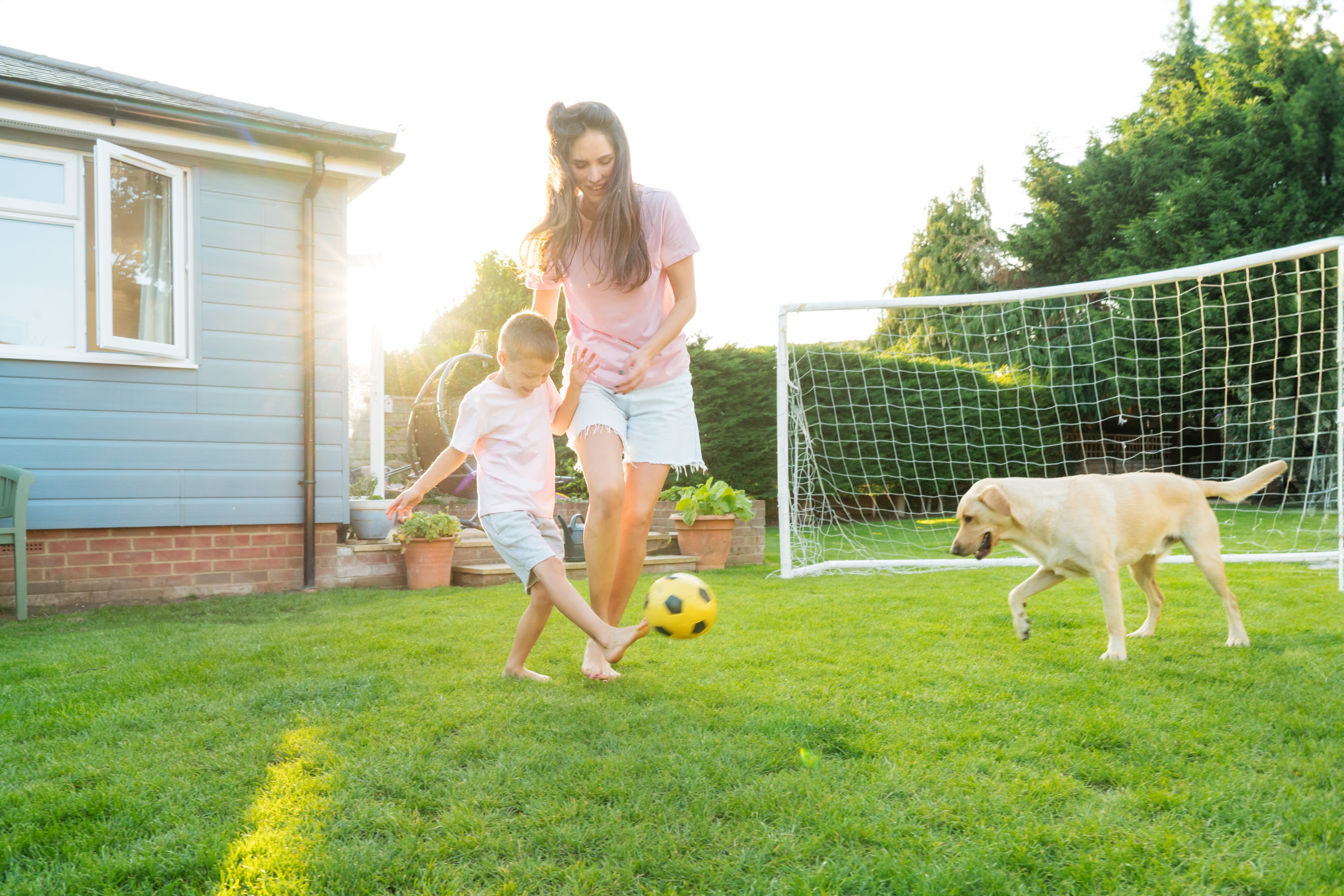 Mother and son plays soccer outside. They are both smiling and a dog is walking nearby. The sun is shining and a home is in the background.