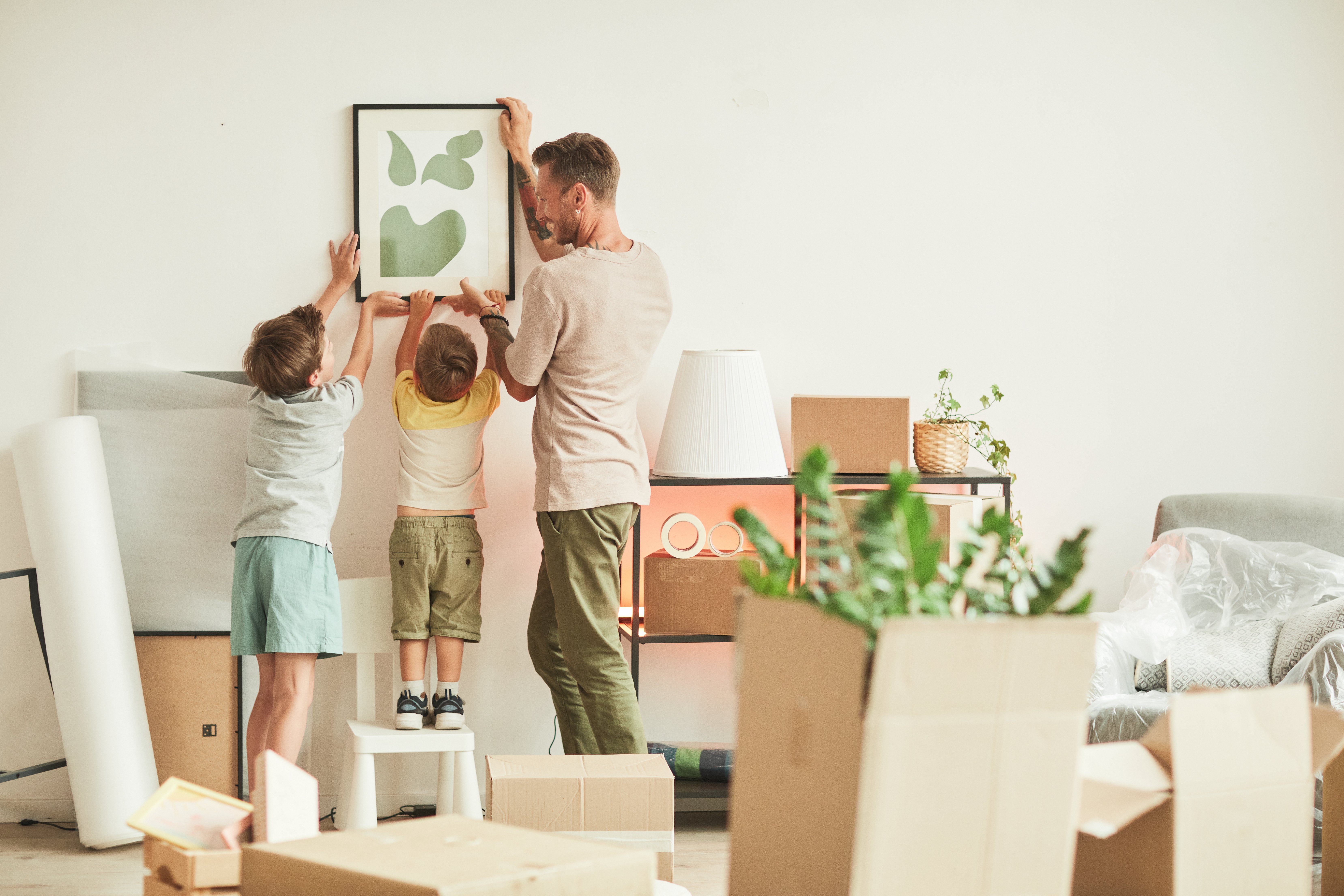 A father and his two young children are hanging a framed abstract art piece on a wall in their new home. The younger child is standing on a stool while the father and older sibling help position the frame. Around them are moving boxes, houseplants, and unpacked home items, indicating they are in the process of settling in. The scene captures a warm, collaborative moment during a family move.