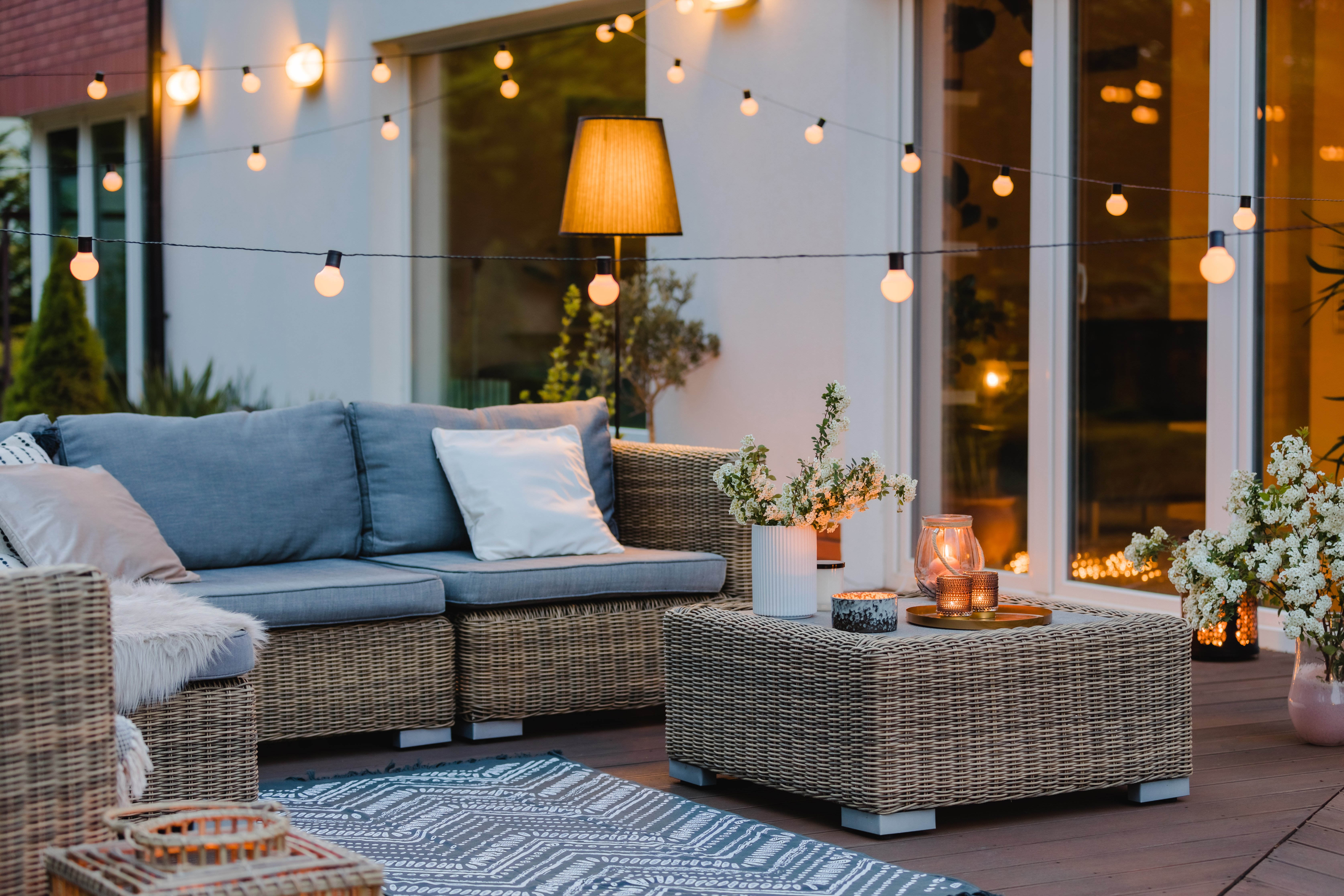 A cozy outdoor patio area in the evening, featuring wicker furniture with gray cushions, a patterned outdoor rug, and string lights hanging above. A coffee table holds a white vase with flowers and several lit candles. A tall floor lamp glows warmly in the background near large glass doors, adding to the inviting ambiance. Additional flower arrangements decorate the space, creating a relaxed and stylish atmosphere.