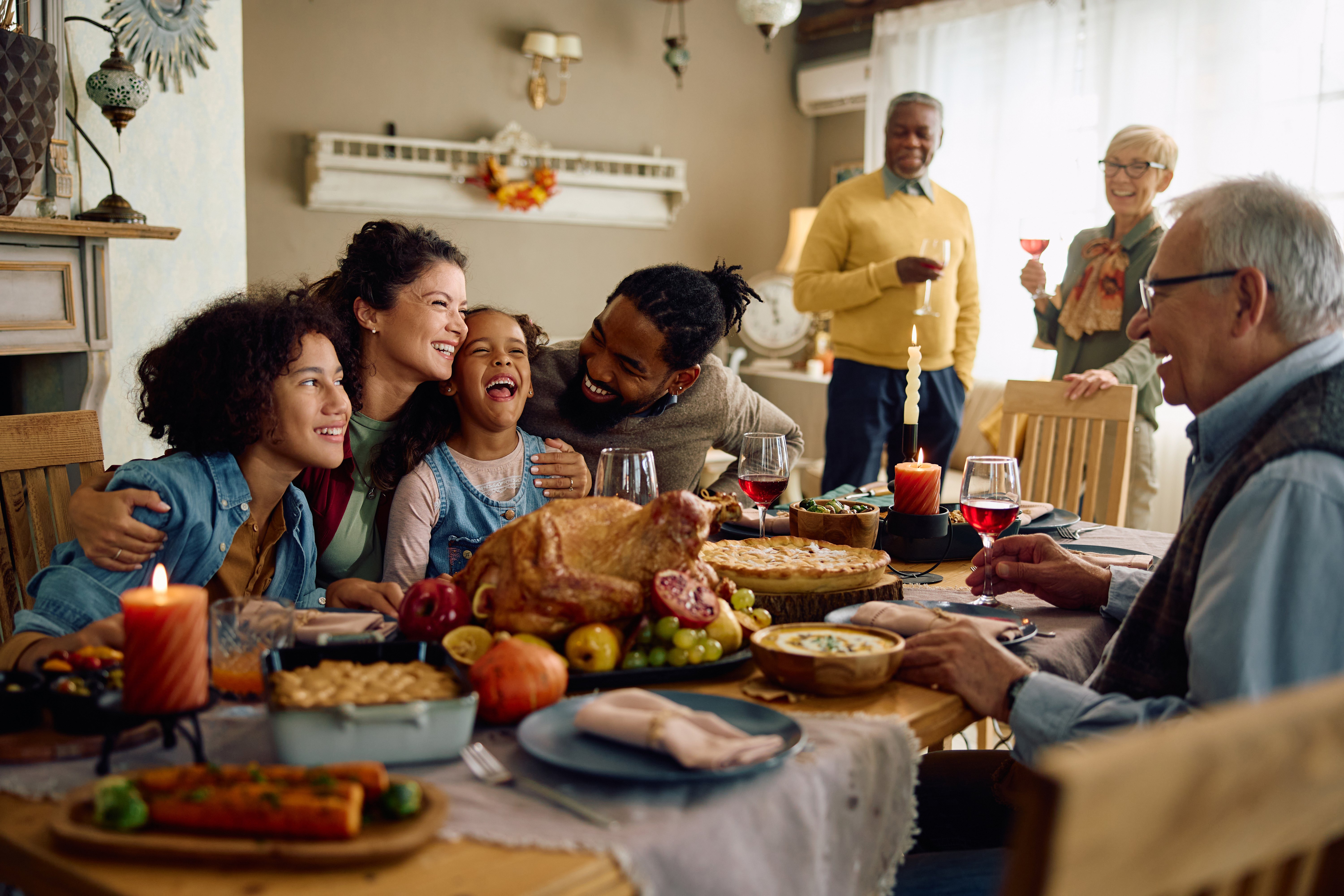 A group of immediate and extended family gather in a living room, some standing, some sitting and smiling with their arms around each other at the dining table. On the dining table, a Thanksgiving meal is set.